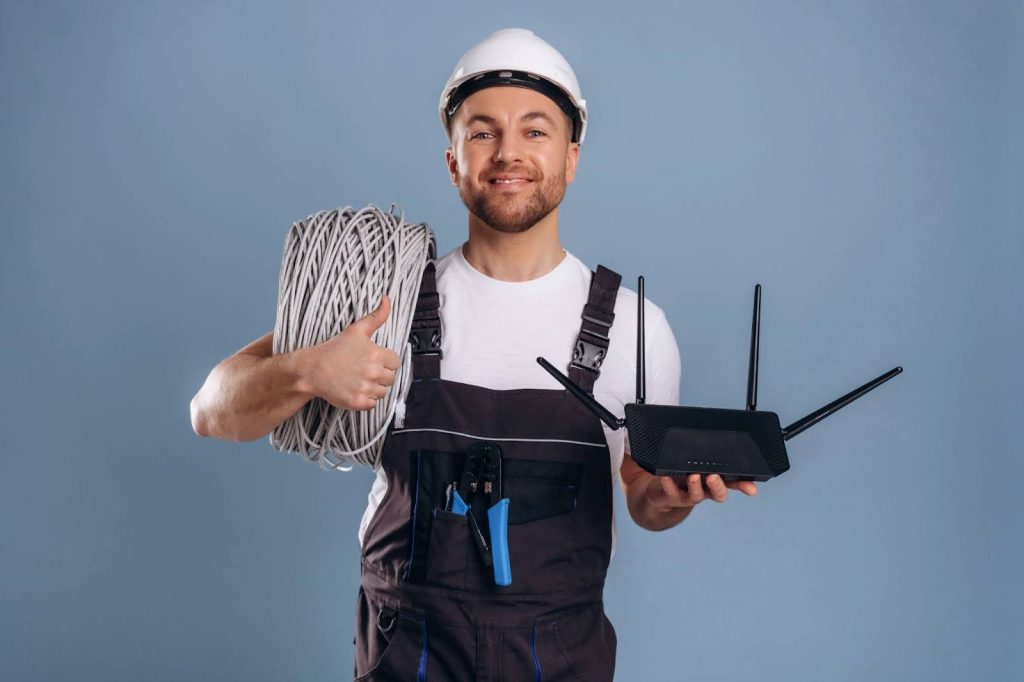 A worker holding a black 4-antenna wireless router and a large roll of grey Ethernet cable, with a crimping tool in his pocket.