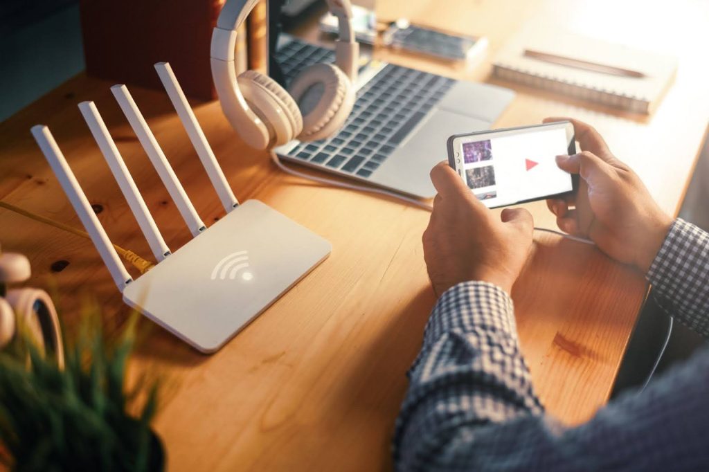 A person holding a smartphone streaming a video while sitting at a wooden desk next to a white, four-antenna Wi-Fi router.